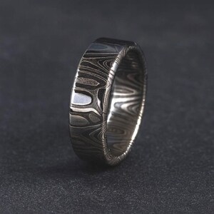 Puede incluir: Primer plano de un anillo plateado con un patr&oacute;n arremolinado &uacute;nico. El anillo tiene un acabado pulido y una banda ancha. El dise&ntilde;o se asemeja a la veta de la madera, con tonos grises claros y oscuros que crean una apariencia texturizada. El anillo est&aacute; sobre un fondo oscuro.