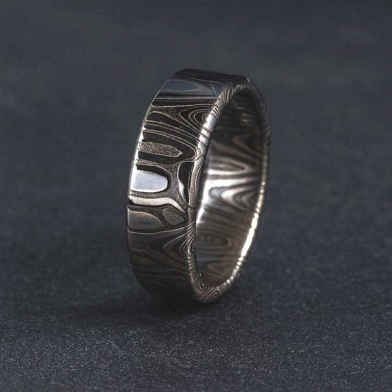 Puede incluir: Primer plano de un anillo plateado con un patr&oacute;n arremolinado &uacute;nico. El anillo tiene un acabado pulido y una banda ancha. El dise&ntilde;o se asemeja a la veta de la madera, con tonos grises claros y oscuros que crean una apariencia texturizada. El anillo est&aacute; sobre un fondo oscuro.