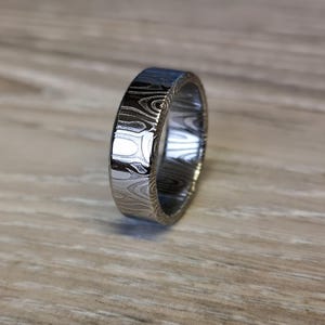 Puede incluir: Un anillo plateado con un patr&oacute;n &uacute;nico de vetas de madera. El anillo tiene un perfil plano y un acabado pulido, que refleja la luz. Esta joya es probablemente para uso diario.
