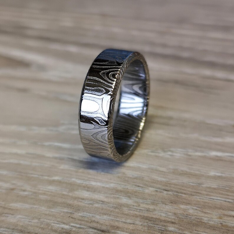 Puede incluir: Un anillo plateado con un patr&oacute;n &uacute;nico de vetas de madera. El anillo tiene un perfil plano y un acabado pulido, que refleja la luz. Esta joya es probablemente para uso diario.