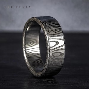Puede incluir: Primer plano de un anillo plateado con un patr&oacute;n arremolinado &uacute;nico. El anillo tiene un perfil plano y una superficie texturizada. El texto "THE FENJA" es visible en la parte superior de la imagen. El anillo est&aacute; sobre un fondo oscuro.