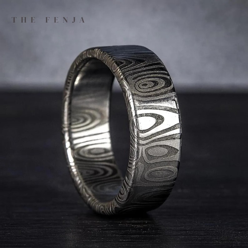 Puede incluir: Primer plano de un anillo plateado con un patr&oacute;n arremolinado &uacute;nico. El anillo tiene un perfil plano y una superficie texturizada. El texto "THE FENJA" es visible en la parte superior de la imagen. El anillo est&aacute; sobre un fondo oscuro.