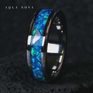 May include: A black ring with a central inlay of blue and green opal. The ring has a polished, reflective surface. The text "AQUA NOVA" is at the top. The opal has a textured appearance. The ring is a piece of jewellery.