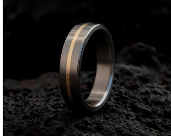 May include: A close-up of a ring with a dark gray band and a central gold stripe. The ring is set against a dark, textured background. The word "AUREUM" is visible at the top.