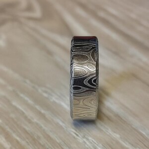 Puede incluir: Primer plano de un anillo estampado. El anillo presenta un dise&ntilde;o &uacute;nico con patrones arremolinados en tonos plateados, negros y un toque de rojo. El anillo est&aacute; hecho de metal y tiene un acabado liso y pulido. El anillo es una joya.