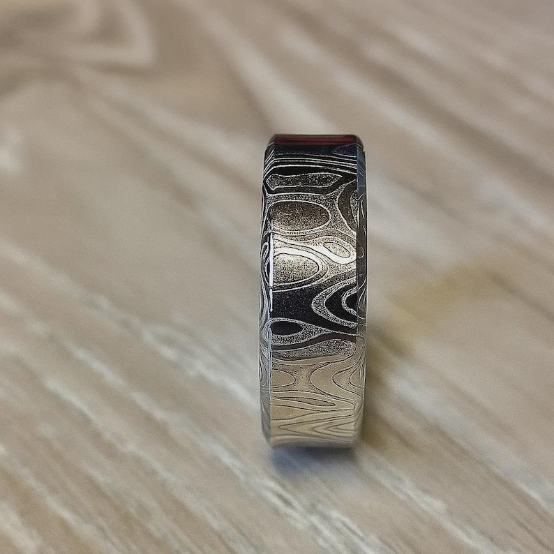Puede incluir: Primer plano de un anillo estampado. El anillo presenta un dise&ntilde;o &uacute;nico con patrones arremolinados en tonos plateados, negros y un toque de rojo. El anillo est&aacute; hecho de metal y tiene un acabado liso y pulido. El anillo es una joya.