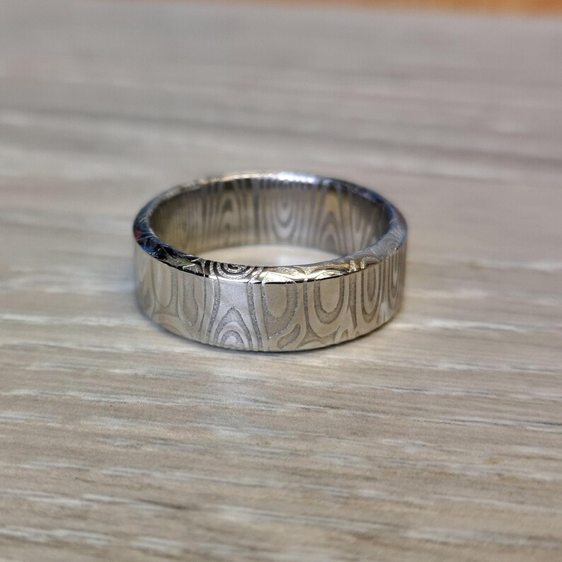 Puede incluir: Un anillo de plata con un patr&oacute;n &uacute;nico de vetas de madera. El anillo es de estilo banda, con una superficie plana y un acabado pulido. El intrincado dise&ntilde;o le da al anillo un aspecto distintivo. El anillo se muestra sobre una superficie de madera de color claro.
