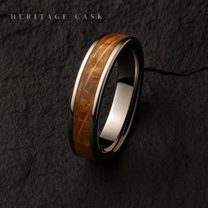 Puede incluir: Primer plano de un anillo con una banda de metal oscuro, una banda interior de metal claro y un centro de vetas de madera. El texto "HERITAGE CASK" está en la parte superior. El anillo está sobre una superficie oscura y texturizada.