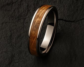 Puede incluir: Primer plano de un anillo con una banda de metal oscuro, una banda interior de metal claro y un centro de vetas de madera. El texto "HERITAGE CASK" está en la parte superior. El anillo está sobre una superficie oscura y texturizada.