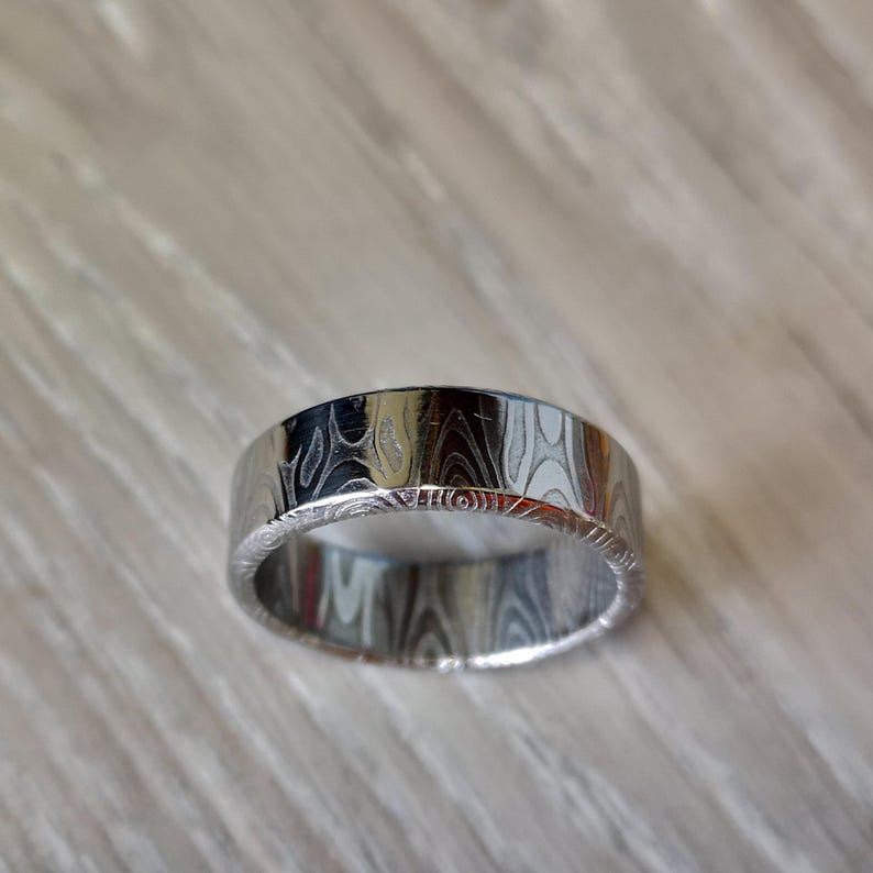 Puede incluir: Un anillo plateado con un patr&oacute;n &uacute;nico de vetas de madera. El anillo tiene una banda plana y ancha con una superficie texturizada. El anillo es probablemente de metal y est&aacute; dise&ntilde;ado para el uso diario.