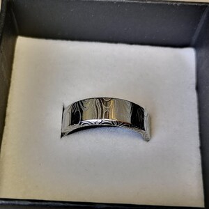 Puede incluir: Un anillo plateado con un dise&ntilde;o estampado, presentado en una caja negra con interior blanco. La superficie del anillo presenta un patr&oacute;n arremolinado y texturizado. El anillo es de estilo banda, adecuado para diversas ocasiones.
