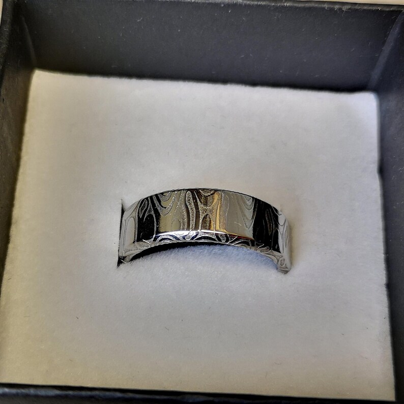 Puede incluir: Un anillo plateado con un dise&ntilde;o estampado, presentado en una caja negra con interior blanco. La superficie del anillo presenta un patr&oacute;n arremolinado y texturizado. El anillo es de estilo banda, adecuado para diversas ocasiones.