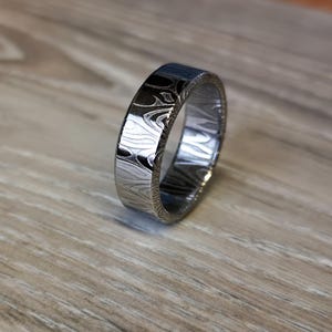 Puede incluir: Un anillo plateado con un dise&ntilde;o &uacute;nico. El anillo tiene una banda plana y una superficie texturizada, creando un efecto visual llamativo. El anillo se muestra sobre una superficie de madera.