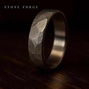 May include: A silver ring with a textured, faceted surface. The ring has a hammered appearance, with the words "STONE FORGE" in the upper left corner. The ring is set against a dark background, highlighting its metallic sheen.