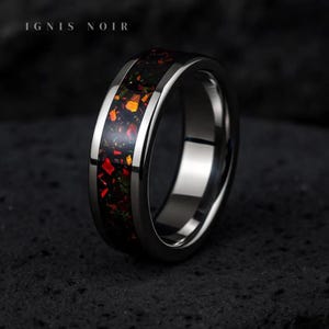 May include: A close-up of a silver ring with a black and red inlay. The ring has a polished finish and the text "IGNIS NOIR" is at the top. The inlay features a mix of red, orange, and green colors.