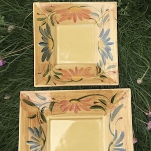 Puede incluir: Tres platos cuadrados de cerámica con un centro amarillo claro y un borde floral decorativo. El borde presenta flores pintadas a mano en tonos rosa, azul y verde. Los platos están dispuestos sobre hierba verde con flores moradas.