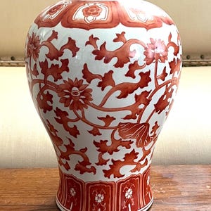 Puede incluir: Un jarrón de cerámica blanca con un patrón floral rojo. El jarrón tiene un cuerpo ancho, un cuello estrecho y un borde acampanado. El diseño floral incluye flores, hojas y vides. El jarrón probablemente se utiliza con fines decorativos.