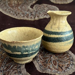 May include: A ceramic sake set featuring a small, round pitcher and a matching cup. The pitcher and cup are beige with dark teal horizontal stripes. The set has a rustic, handmade appearance, suggesting traditional Japanese craftsmanship.