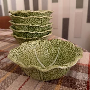 Peut inclure: Une pile de six bols en céramique verte, chacun présentant une surface texturée imitant l'apparence d'une feuille de chou avec des nervures blanches saillantes et des bords ondulés. Les bols ont une finition émaillée brillante et nuancée.