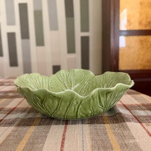 Może przedstawiać: Jasnozielona ceramiczna miska w kształcie liścia kapusty. Miska ma realistyczną, teksturowaną powierzchnię z wytłoczonymi żyłkami i pofalowanymi krawędziami, które naśladują wygląd naturalnych liści, tworząc wyrazisty, organiczny i zaokrąglony kształt.