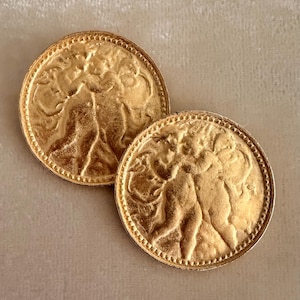 May include: Two gold-colored, round earrings with a detailed relief design. The earrings feature a textured border and a central image of two figures. The earrings are displayed on a neutral-toned surface.
