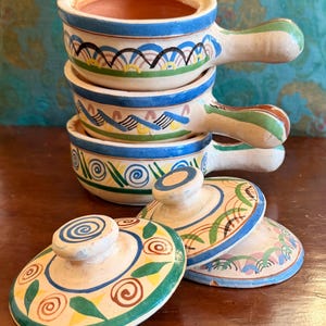 Puede incluir: Cuencos de sopa de cerámica apilados con tapas. Los cuencos blancos tienen diseños pintados en azul, verde, amarillo y marrón. Cada cuenco tiene un asa. Las tapas tienen un diseño de remolino.