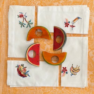Pode incluir: Guardanapos de linho branco com bordados de frutas e flores, combinados com argolas de guardanapo de madeira em forma de melancia e cantalupo. Os guardanapos apresentam bordados coloridos de um coquetel, bananas e uma cesta de frutas.
