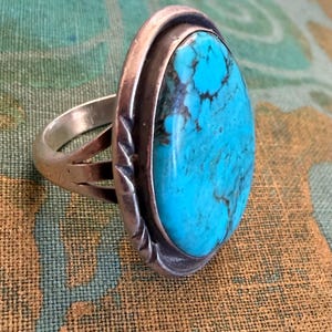 Puede incluir: Un anillo de plata con una gran piedra turquesa ovalada. La piedra azul brillante tiene vetas oscuras y está engastada en un bisel de plata. El anillo tiene un diseño de caña dividida con detalles decorativos en los laterales.