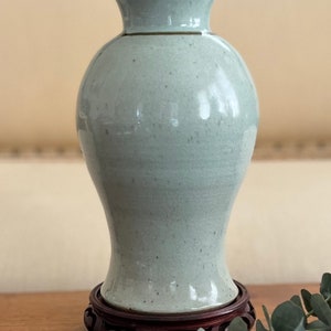 May include: A pale green ceramic vase with a flared rim and a bulbous body, set on a dark wooden stand. The vase has a smooth, glossy finish with subtle speckling. Decorative home decor.