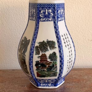 Puede incluir: Jarrón de cerámica blanco y azul con forma hexagonal. El jarrón presenta un paisaje con una pagoda y montañas, enmarcado por patrones florales y geométricos azules. Se ven caracteres chinos en el lateral.