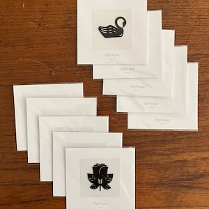 Può includere: Una collezione di biglietti Diptyque Paris con disegni a silhouette nere di un cigno e una rosa. I biglietti sono su sfondi bianchi e sono esposti su una superficie di legno. Il logo Diptyque Paris è stampato sui biglietti.