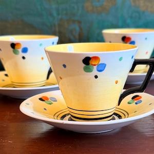 May include: A set of three vintage teacups and saucers. The cups have a yellow interior and a white exterior with colourful polka dots. Black handles and black stripes on the saucers. The teacups are on a wooden surface.