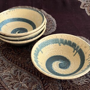 May include: A set of four small, handmade ceramic bowls with a cream-colored base and a blue spiral design in the center. One bowl is separate, showcasing the design. The bowls are stacked on a patterned surface.