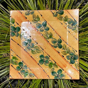 Puede incluir: Plato cuadrado de cerámica con fondo amarillo y rayas diagonales naranjas. El plato está decorado con motivos florales y de hojas verdes, acentuados con líneas negras y puntos blancos. Es un objeto decorativo.