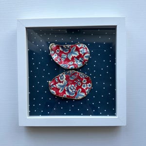 May include: Two decorative oyster shells with red floral patterns and gold edges are displayed in a white framed shadow box. The background is a dark blue fabric with white star patterns. The shells are arranged vertically.