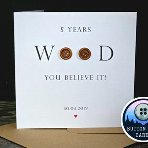 May include: A white card with the text "5 YEARS WOOD YOU BELIEVE IT!" and the date "30.03.2019". The word "WOOD" has two brown buttons in place of the "O"s. A red heart is below the date. The card is on a brown surface. A logo for "BUTTON LOON CARDS" is in the lower right corner.