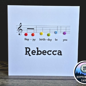 May include: A white card with a musical stave and colourful buttons spelling out "Happy Birthday to You". The name "Rebecca" is written below the music. The card has a logo for "Button Loon Cards" in the bottom right corner.