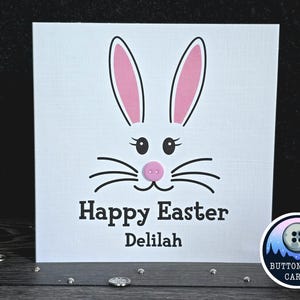 May include: A white card with a pink bunny face and the text "Happy Easter Delilah". The bunny has black whiskers and a pink button nose.