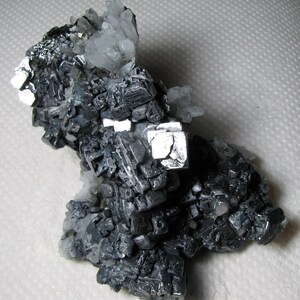 May include: A cluster of gray and silver cubic crystals with a metallic sheen. The crystals are arranged in a random pattern and are surrounded by a white background.