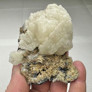 May include: A natural mineral specimen. A large, pale yellow crystalline formation sits on a rough, textured base. The base has a mix of tan, grey, and black elements. The overall structure is irregular and organic in shape.