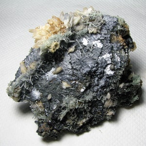 May include: A cluster of gray, metallic, and white crystals on a dark gray rock. The crystals are arranged in a random pattern and have a variety of shapes and sizes.