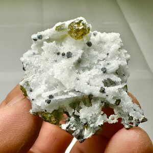 May include: A close-up of a mineral specimen featuring a cluster of white, crystalline formations with small, dark gray and green inclusions. A prominent, golden-yellow crystal sits atop the cluster. The specimen is held in a hand.
