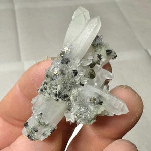 Könnte beinhalten: Nahaufnahme eines Quarzkristallclusters mit transparenten Quarzkristallspitzen und schwarzen, goldenen und grünen Mineraleinschlüssen. Die natürliche Formation zeigt eine komplexe Struktur. Die Quarzkristalle sind gut definiert und optisch ansprechend.
