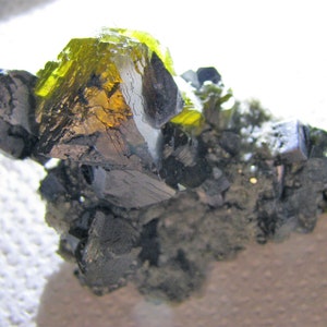 May include: A cluster of dark gray crystals with a large, bright green and yellow crystal on top.