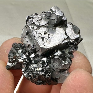 May include: A cluster of metallic, silver-gray mineral crystals. The crystals have a geometric, angular structure, with sharp edges and a reflective surface. The overall shape is irregular and textured, with a mix of large and small crystal formations.