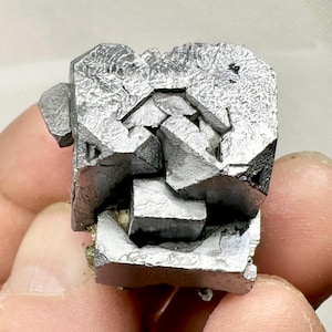 May include: A metallic silver galena mineral specimen featuring a cluster of cubic crystals with sharp, geometric edges and a reflective, textured surface. The mineral displays a complex, multi-faceted structure with deep crevices between the cubes.