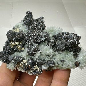 Può includere: Un gruppo di minerali con cristalli grigio scuro e metallici e scaglie dorate di pirite su un letto di cristalli traslucidi verde pallido. La formazione minerale è tenuta in mano.