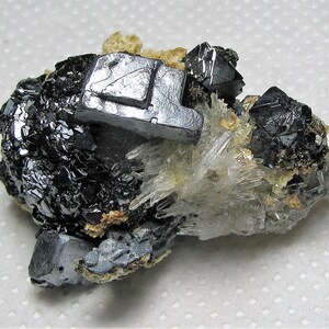May include: A cluster of black and silver crystals on a white background. The crystals are arranged in a random pattern and some are larger than others. The crystals are shiny and have a metallic luster.
