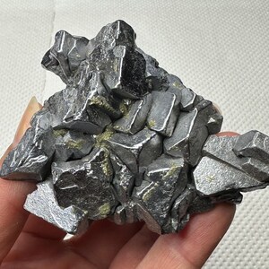 May include: A cluster of metallic gray mineral crystals with sharp, angular facets. The crystals have a reflective surface and are arranged in a complex, irregular formation. Some areas show hints of a golden hue.