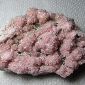 Könnte beinhalten: Ein Cluster aus rosa Rosenquarzkristallen auf einer grauen Matrix. Die Kristalle sind klein und haben eine raue, strukturierte Oberfläche.